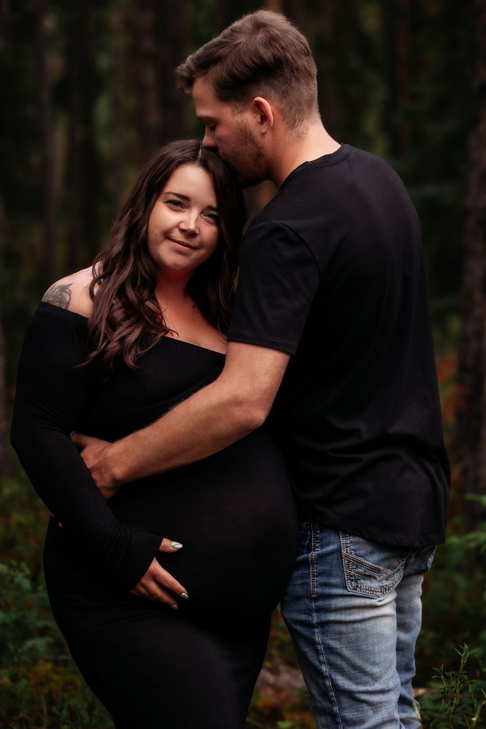 Loving maternity portrait of couple in casual wear embracing in forest setting.