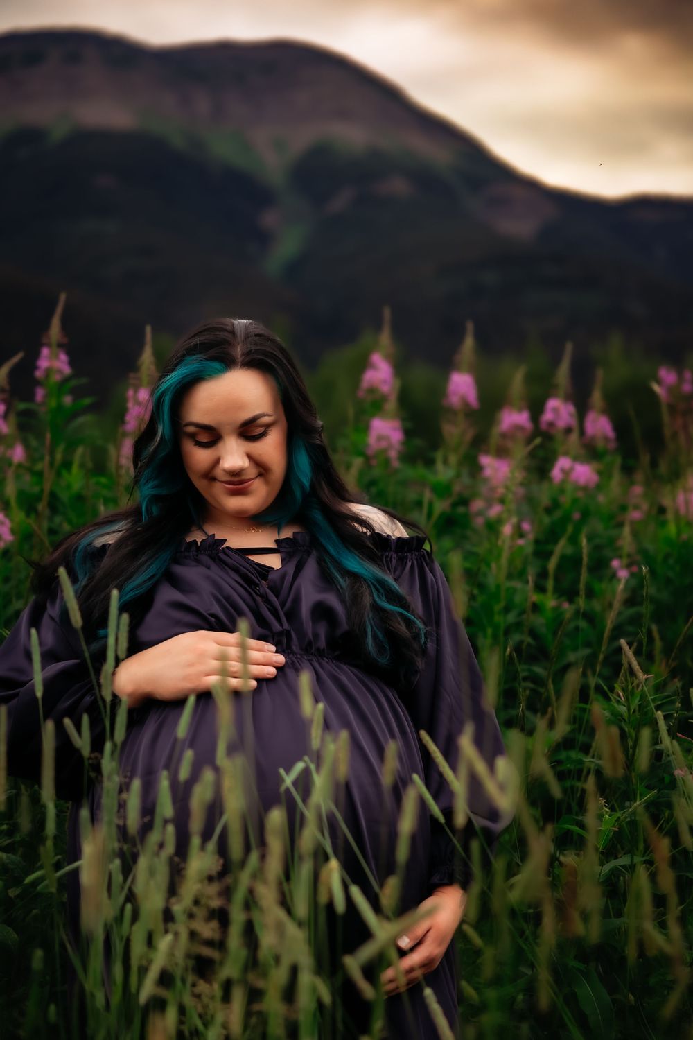 A pregnant woman in a navy dress stands among pink wildflowers with mountains in the background at sunset.