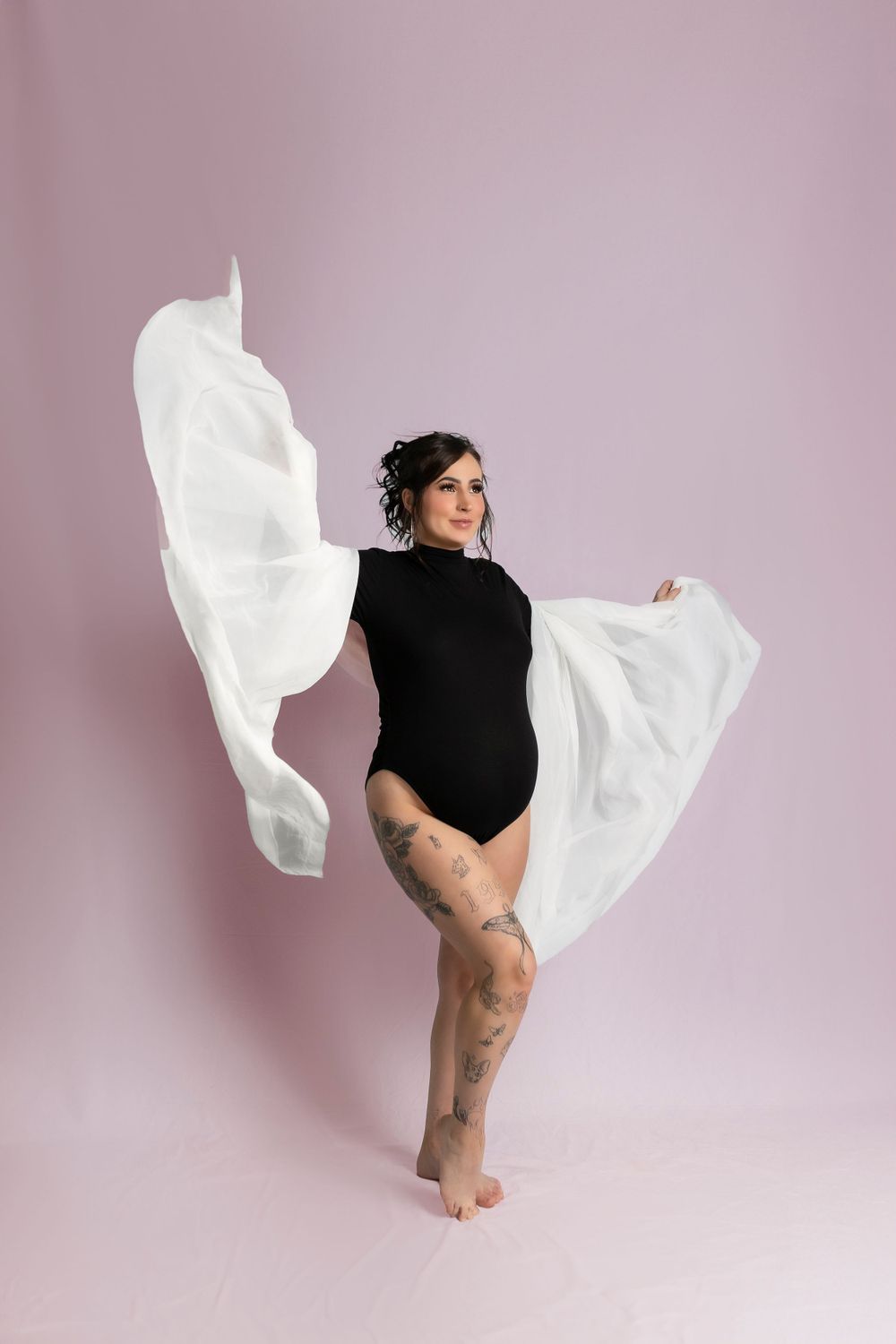 Artistic maternity photo with flowing white fabric against pink backdrop as dancer poses in black leotard.