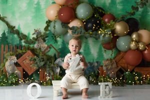 Rustic woodland first birthday setup with green backdrop and earth-toned balloon garland.