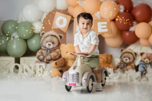 First birthday setup with vintage ride-on car, teddy bear decorations, and green and peach balloon arch.