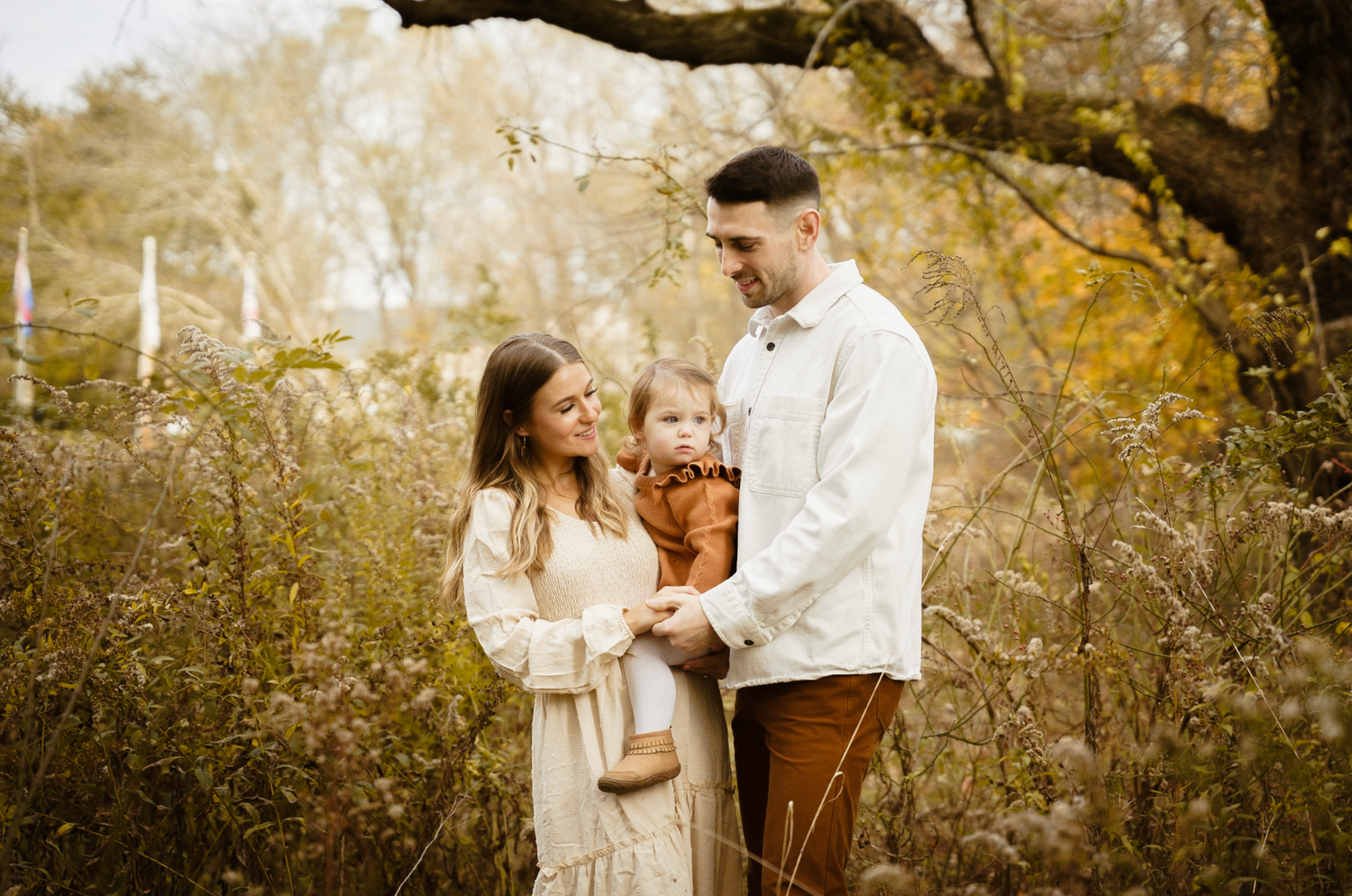 a young couple holding their toddler daughter and looking at her lovingly as they pose in a field during the fall