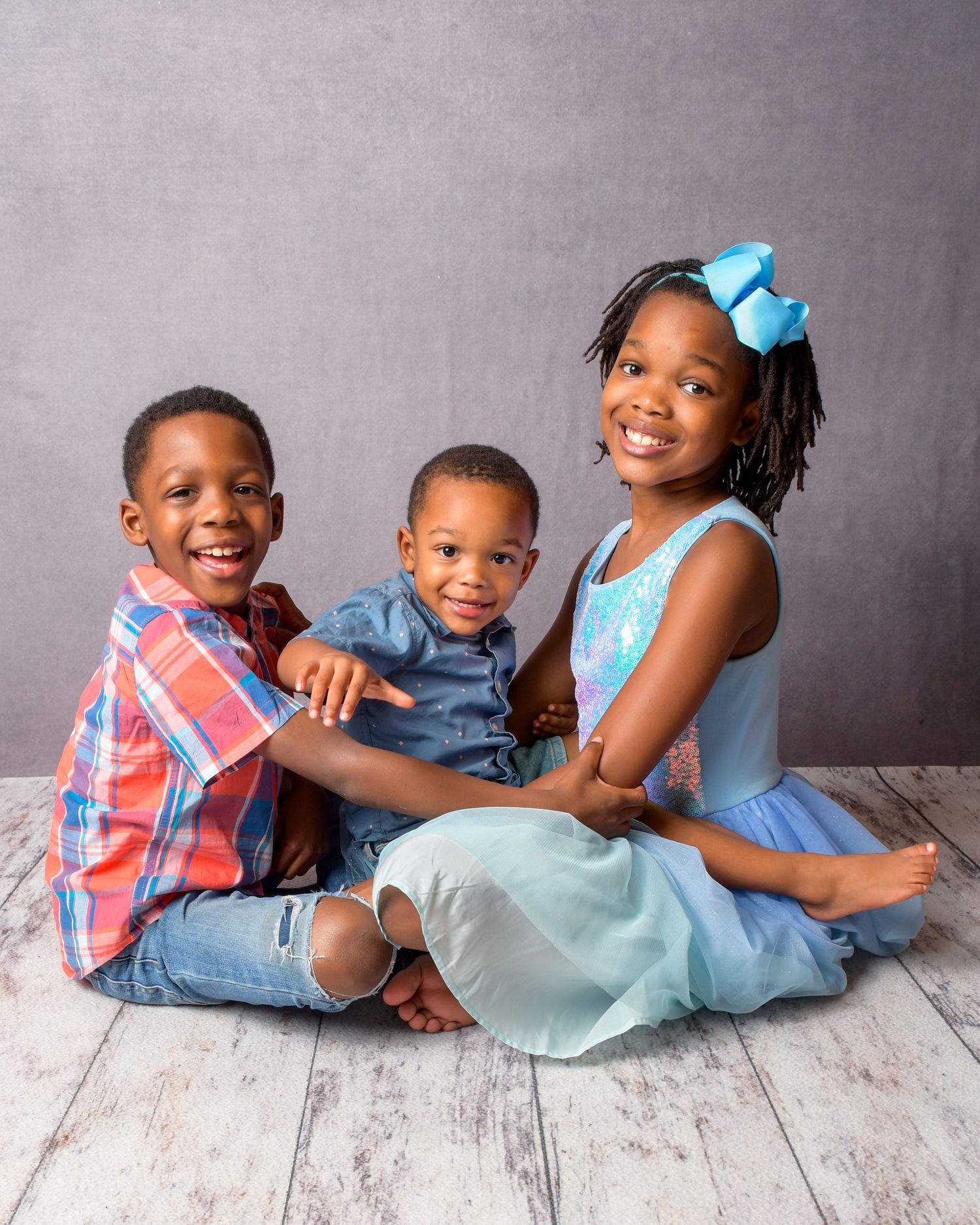 Three siblings in casual attire posing together on wooden floor against grey background.