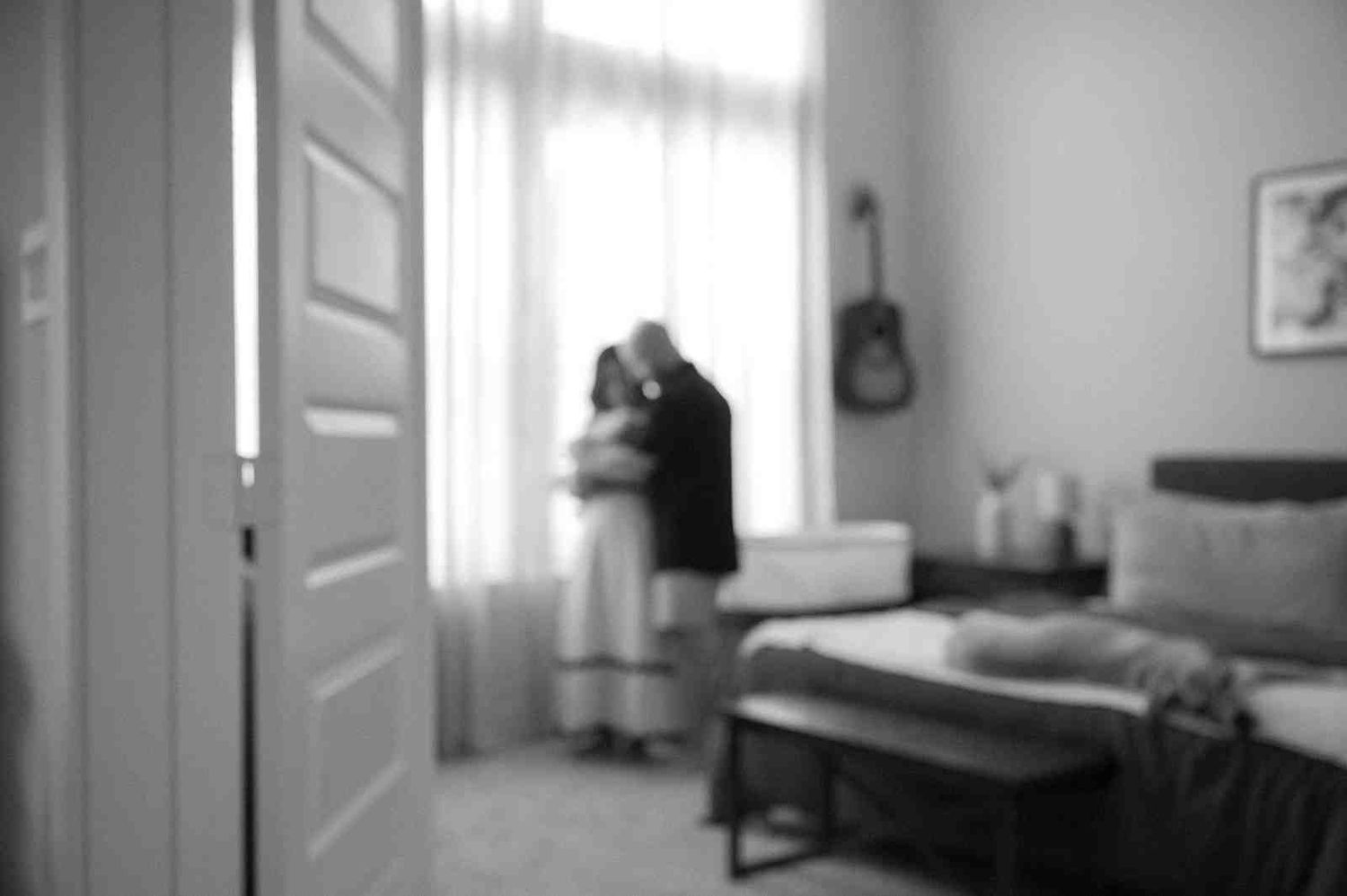 Blurry black and white photo of a bedroom interior with silhouettes near a window and a guitar hanging on the wall.
