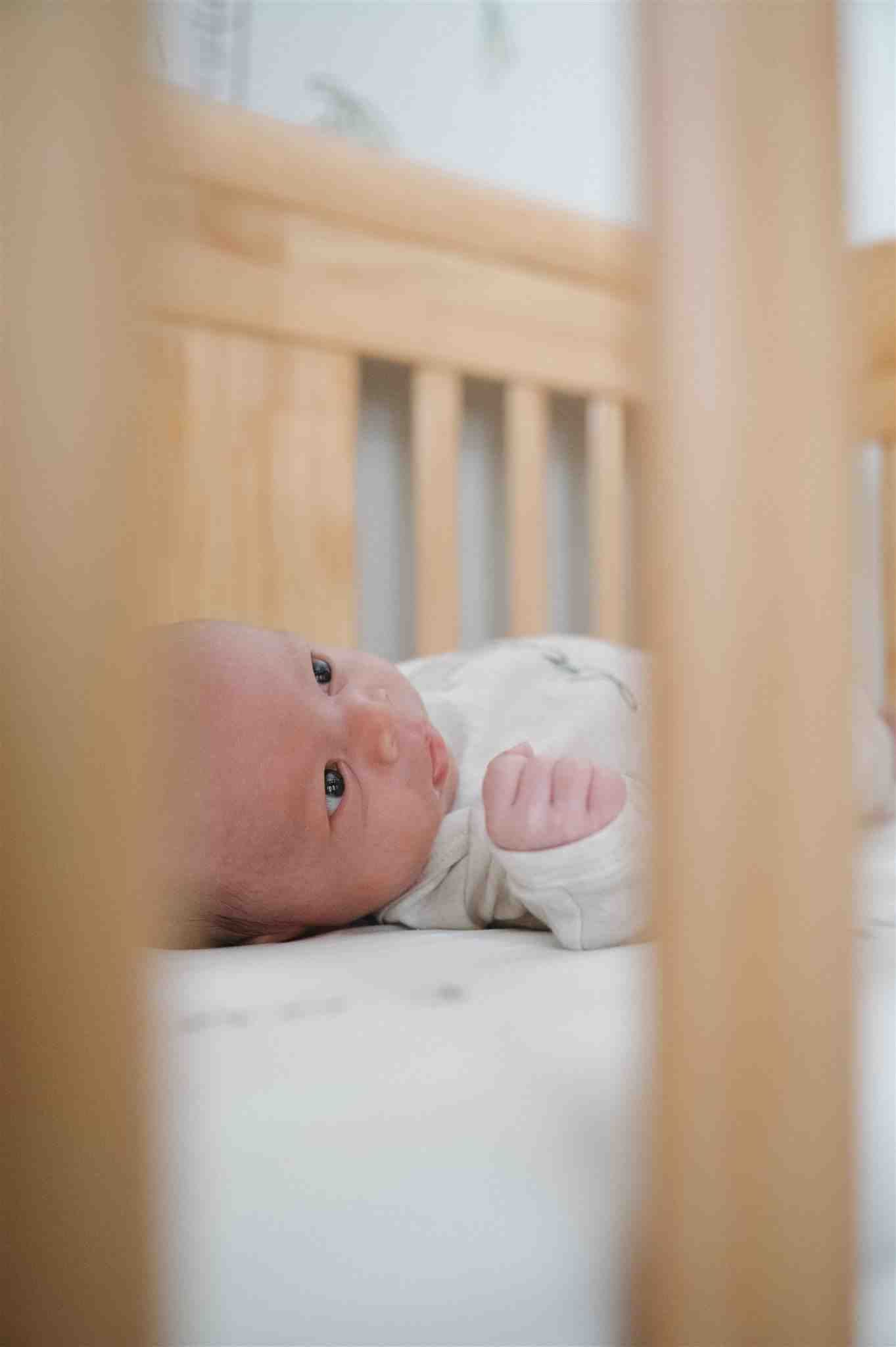 A baby lying in a wooden crib viewed through the slats with soft natural lighting.