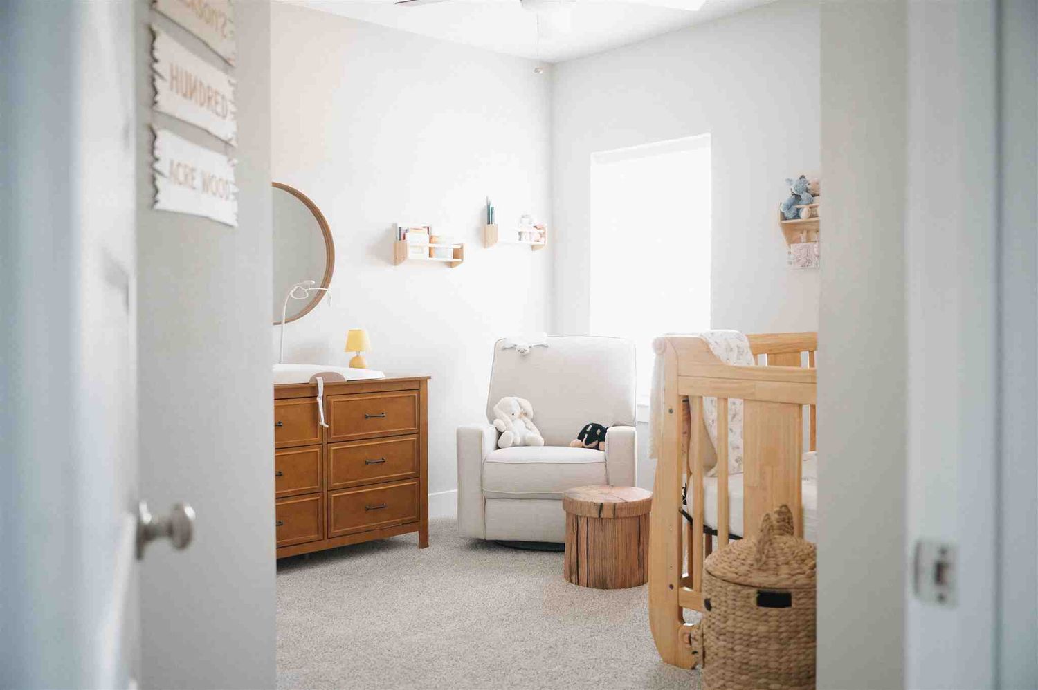A bright modern nursery with wooden furniture, white armchair, and natural light streaming through the window.