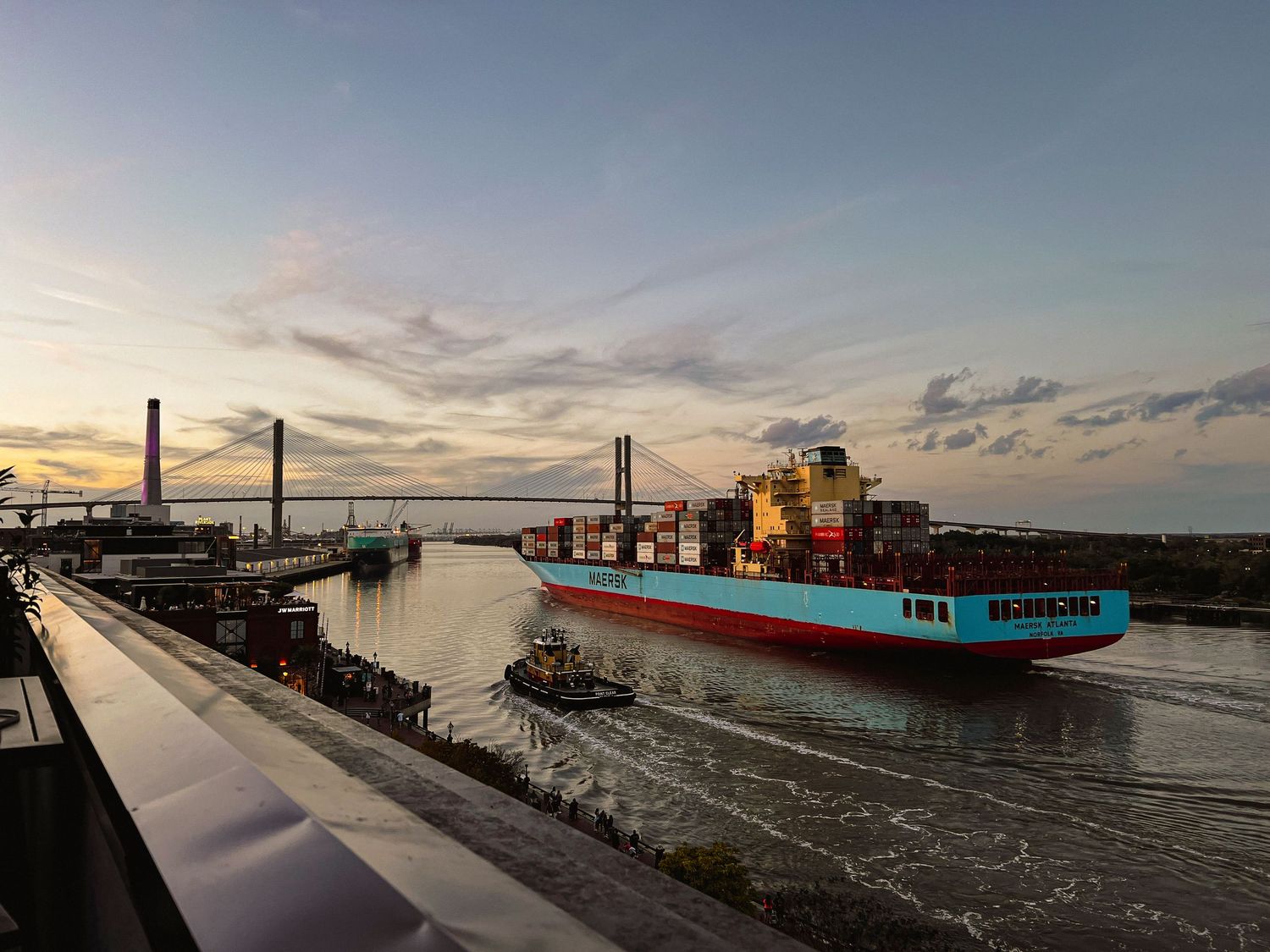 Cargo ship passing under bridge during sunset at industrial port.