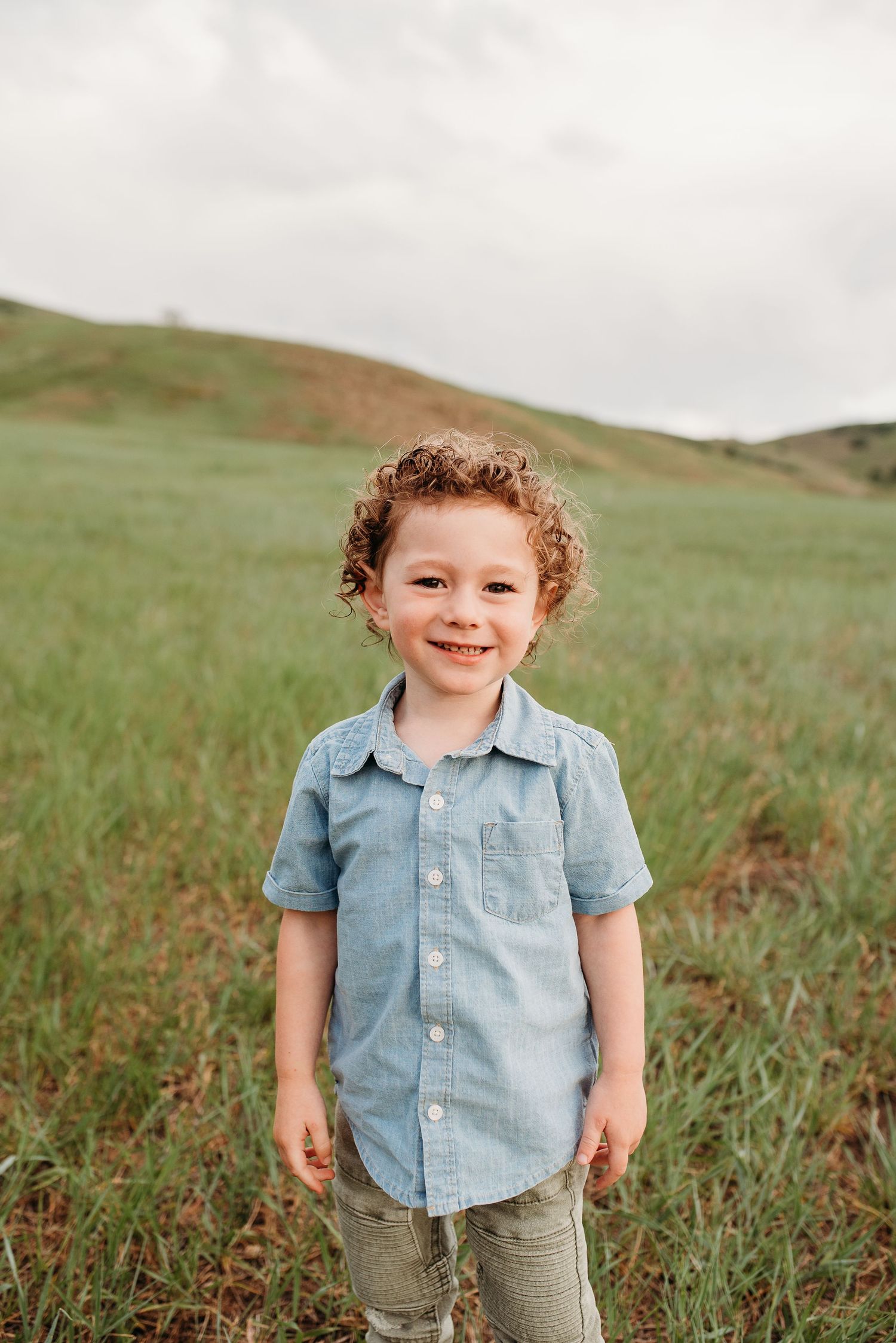 Child in denim shirt smiling in front of rolling green hills during outdoor photo session.