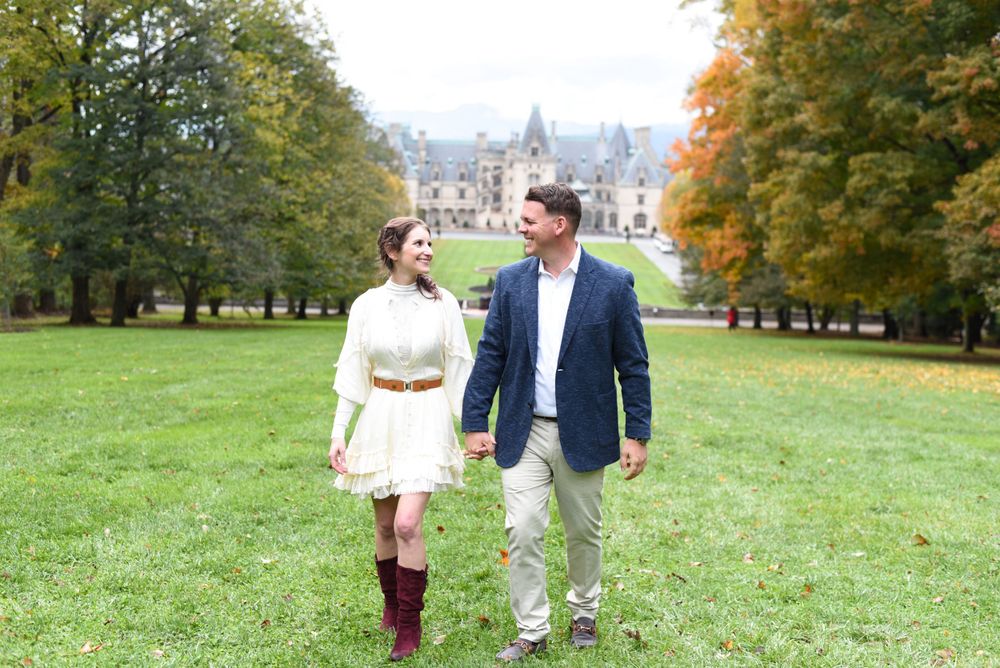 Couple walking hand in hand across castle grounds with fall foliage in background.
