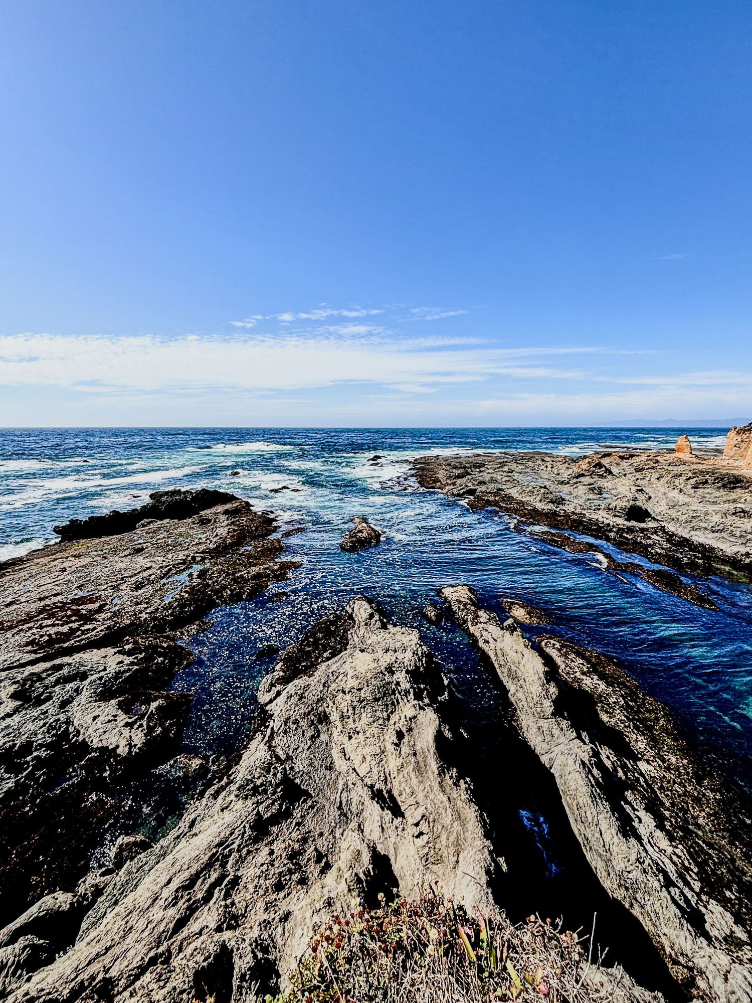 Dramatic coastal scene with rocky outcrops and crashing waves under bright blue sky.