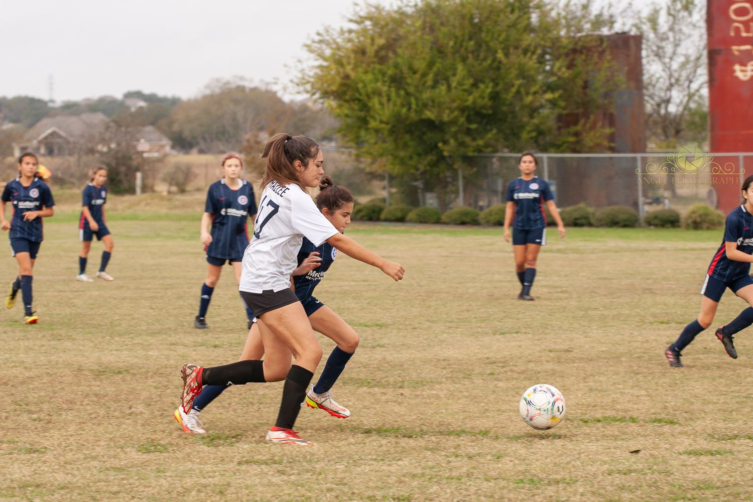Soccer player dribbles ball during match with opponents in navy uniforms in background.