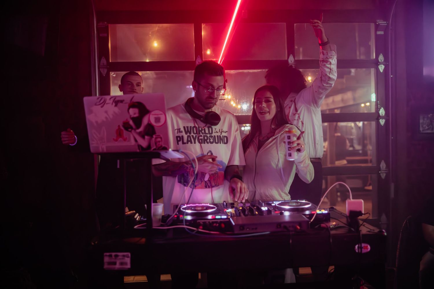 DJ performing at a nightclub with red lighting and mixing equipment on a turntable setup.