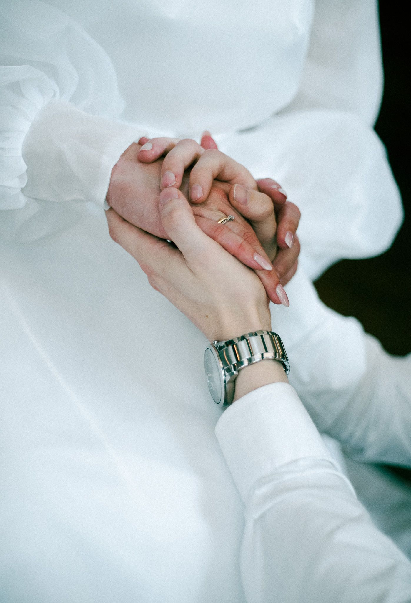 Two hands clasped together in a tender embrace wearing white sleeves and a silver watch.