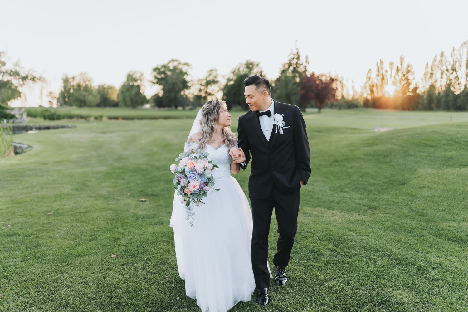 Wedding couple walking on golf course with purple floral bouquet at sunset.