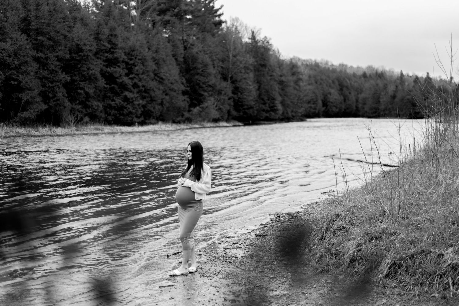 Black and white maternity photo by a riverside with pine trees in the background.
