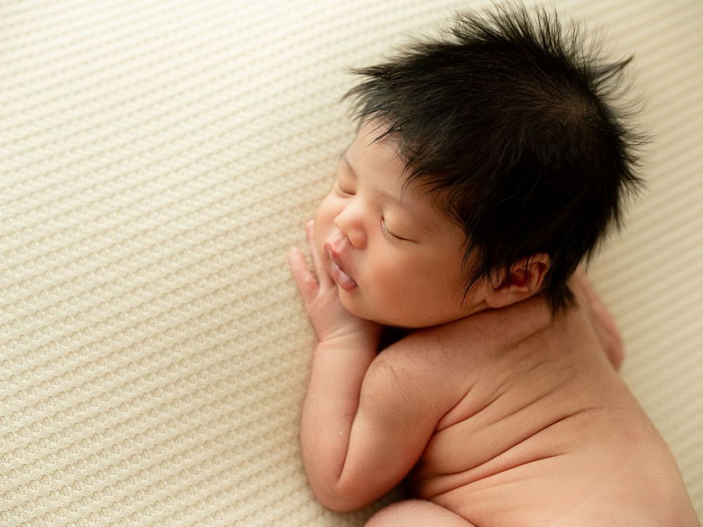 A sleeping baby lies on white bedding with their hands raised near their face.