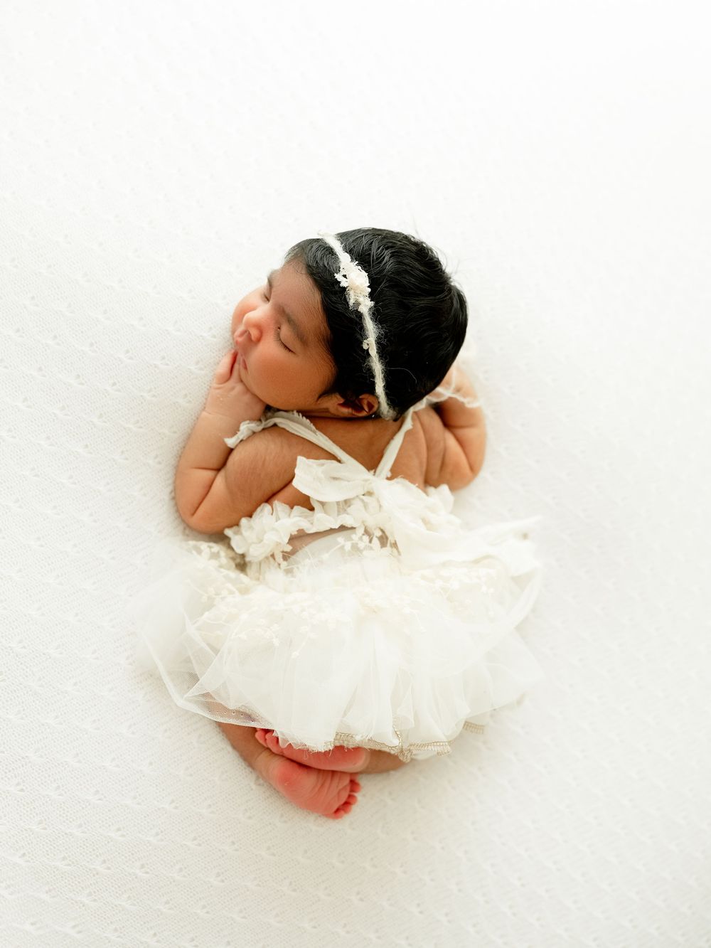 A sleeping baby wearing a white tulle dress and delicate headband lies peacefully on a light background.