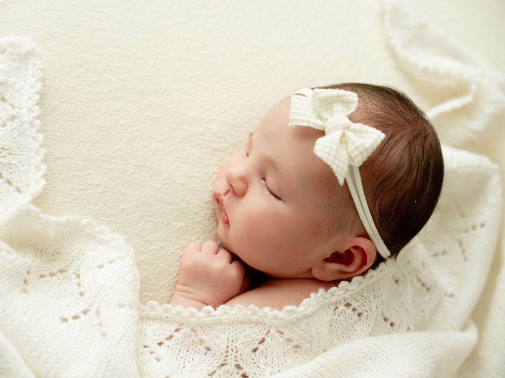 A sleeping baby wearing a white lace headband rests peacefully on soft white bedding.