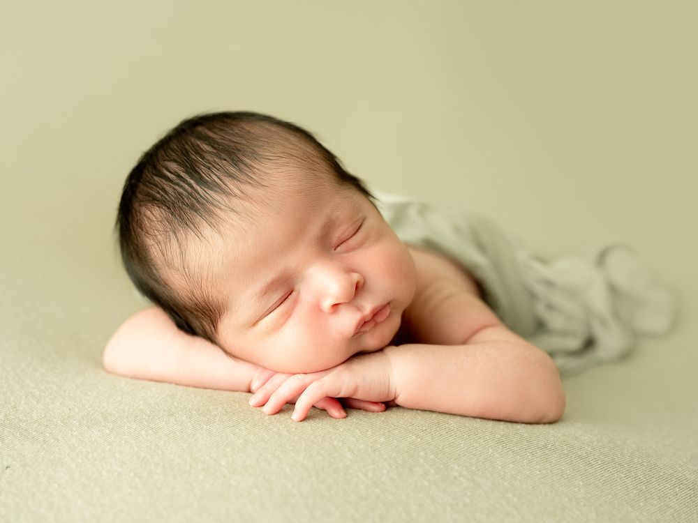 Peaceful sleeping newborn rests with head on arms against soft beige backdrop in gentle lighting.