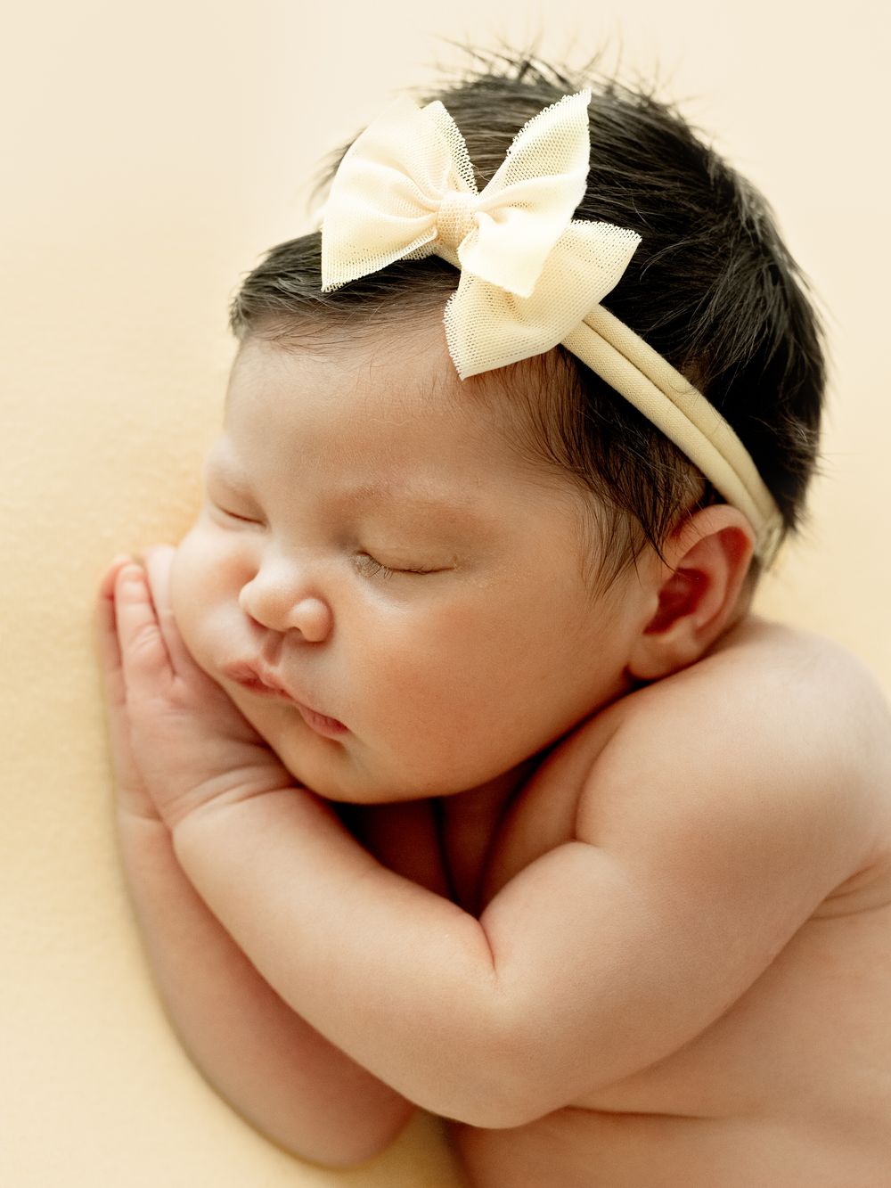 Sleeping newborn wearing cream bow headband on light background showing peaceful expression.