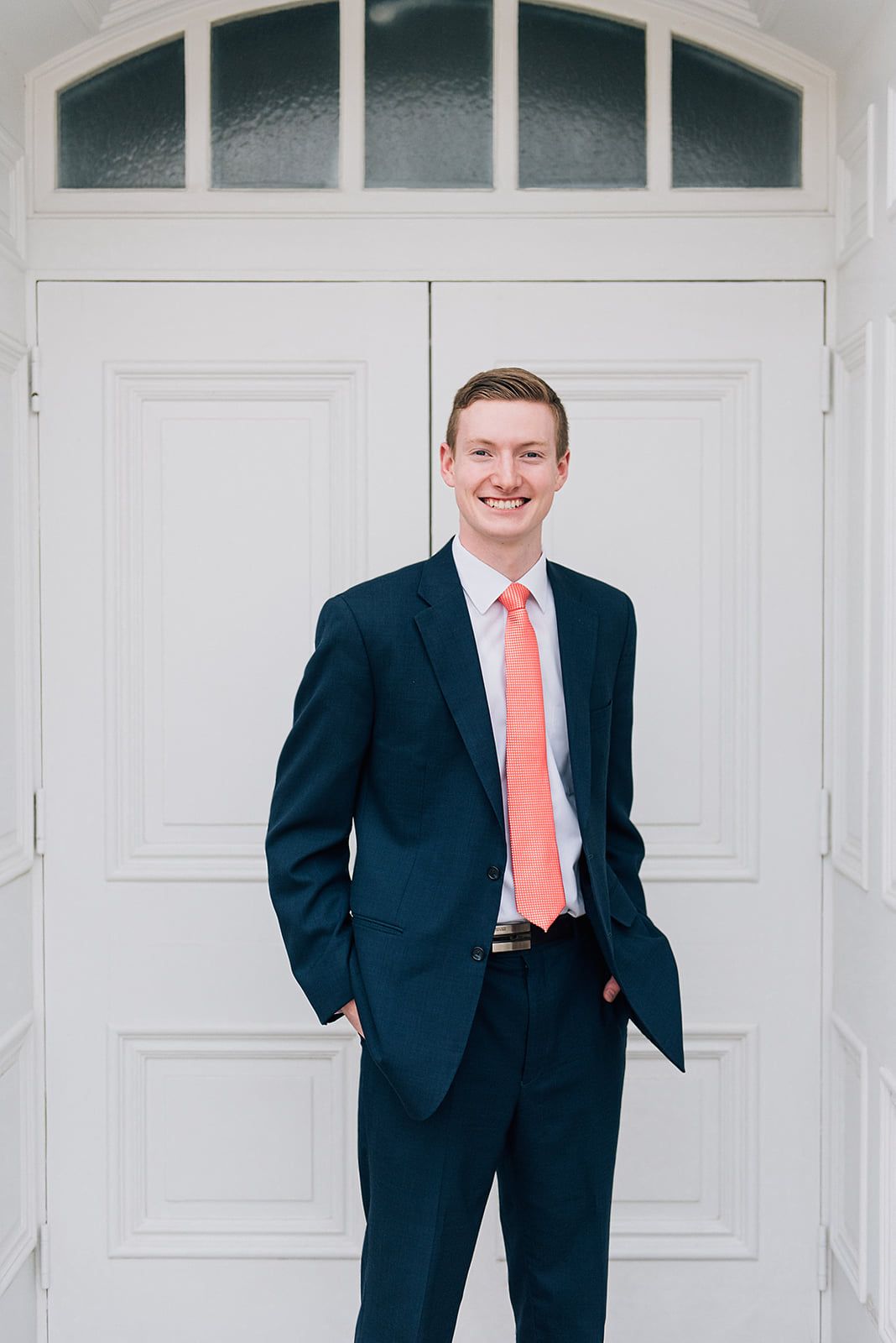 Professional portrait in front of white doors wearing a navy suit with coral tie.