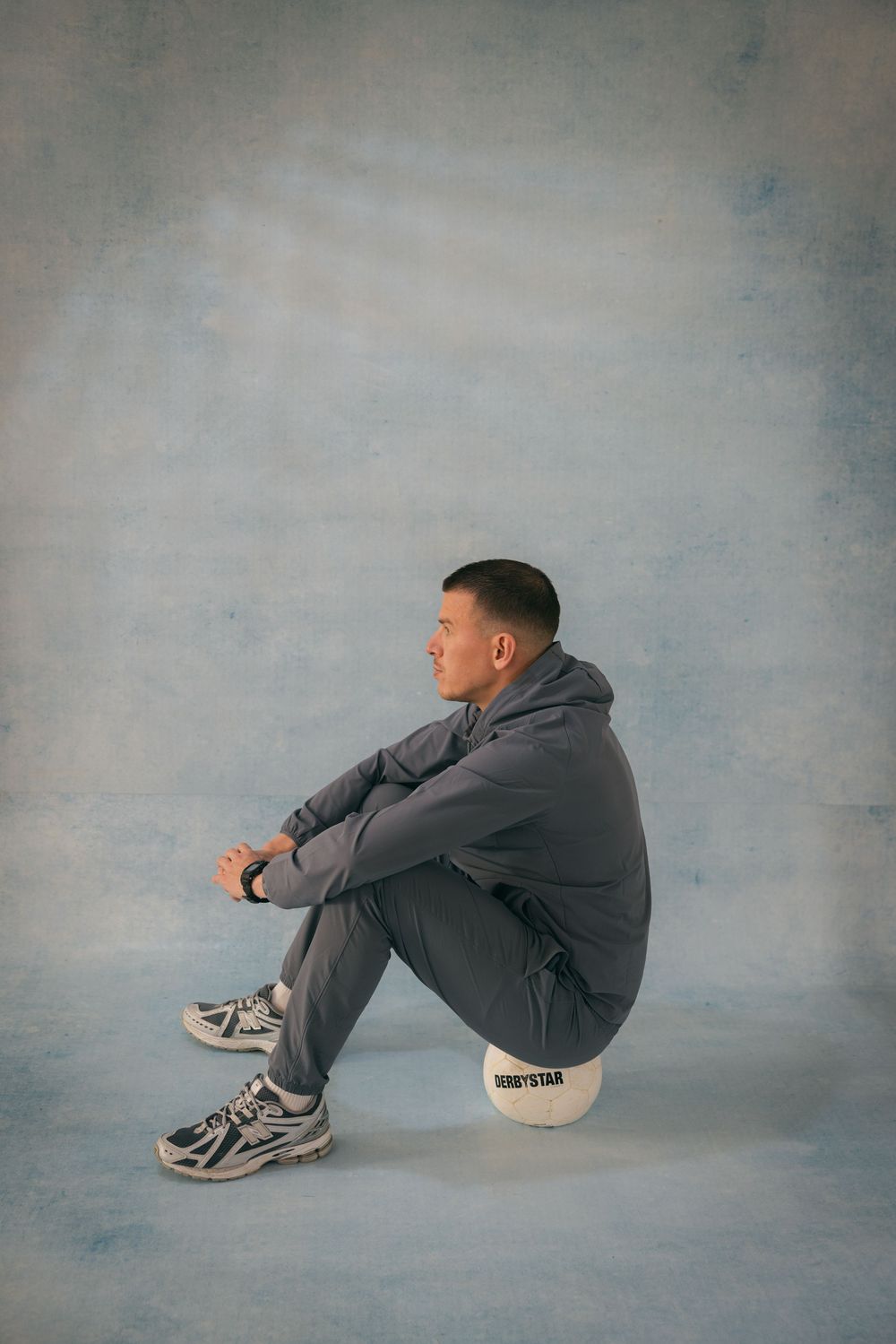 Man in gray tracksuit sitting on a ball wearing sneakers against a blue-gray studio background.