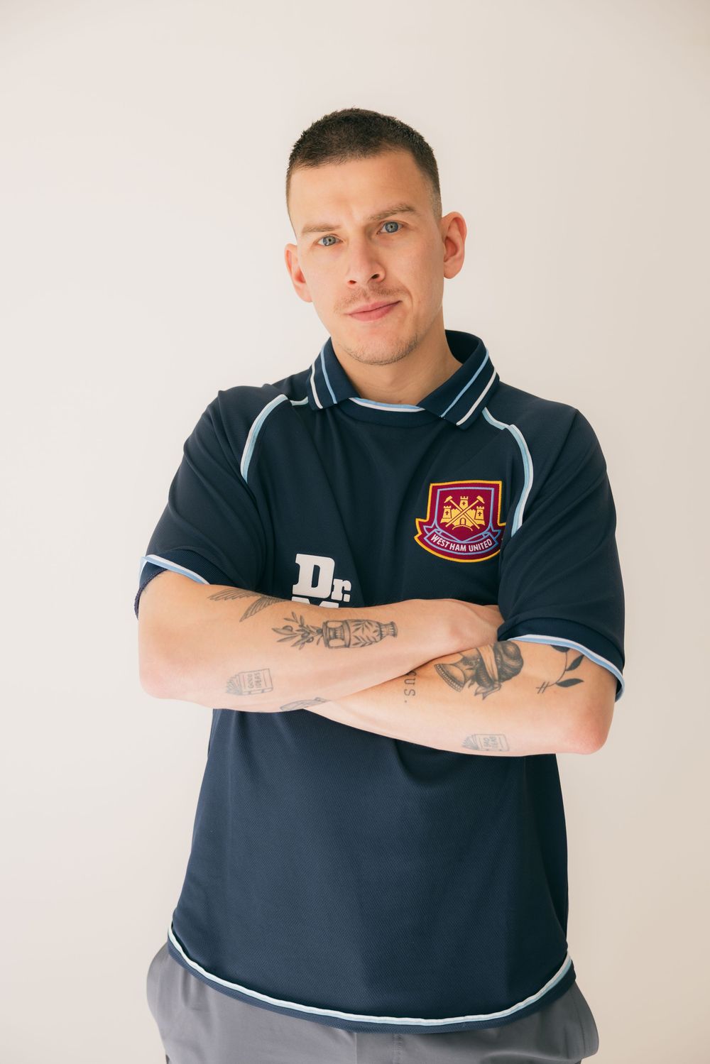 Man with tattoos wearing a navy blue soccer jersey with arms crossed, posing against a white background.