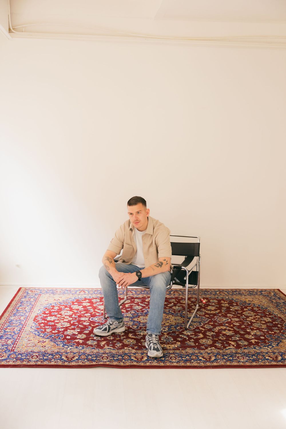 Man in beige jacket and jeans sitting on modern chair atop a red Persian rug against white wall.