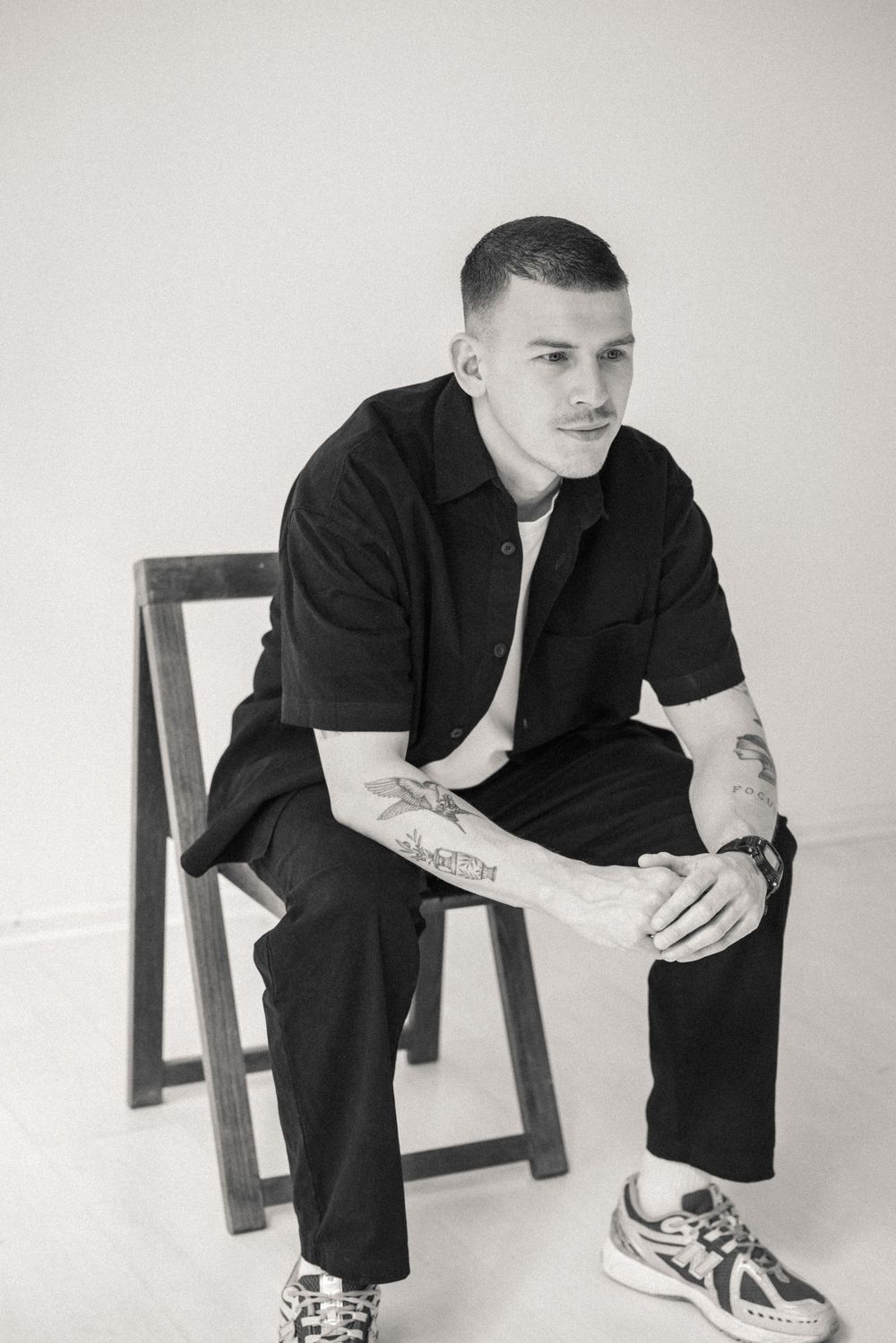 Black and white photo of a tattooed man in black outfit sitting on wooden chair wearing sneakers.