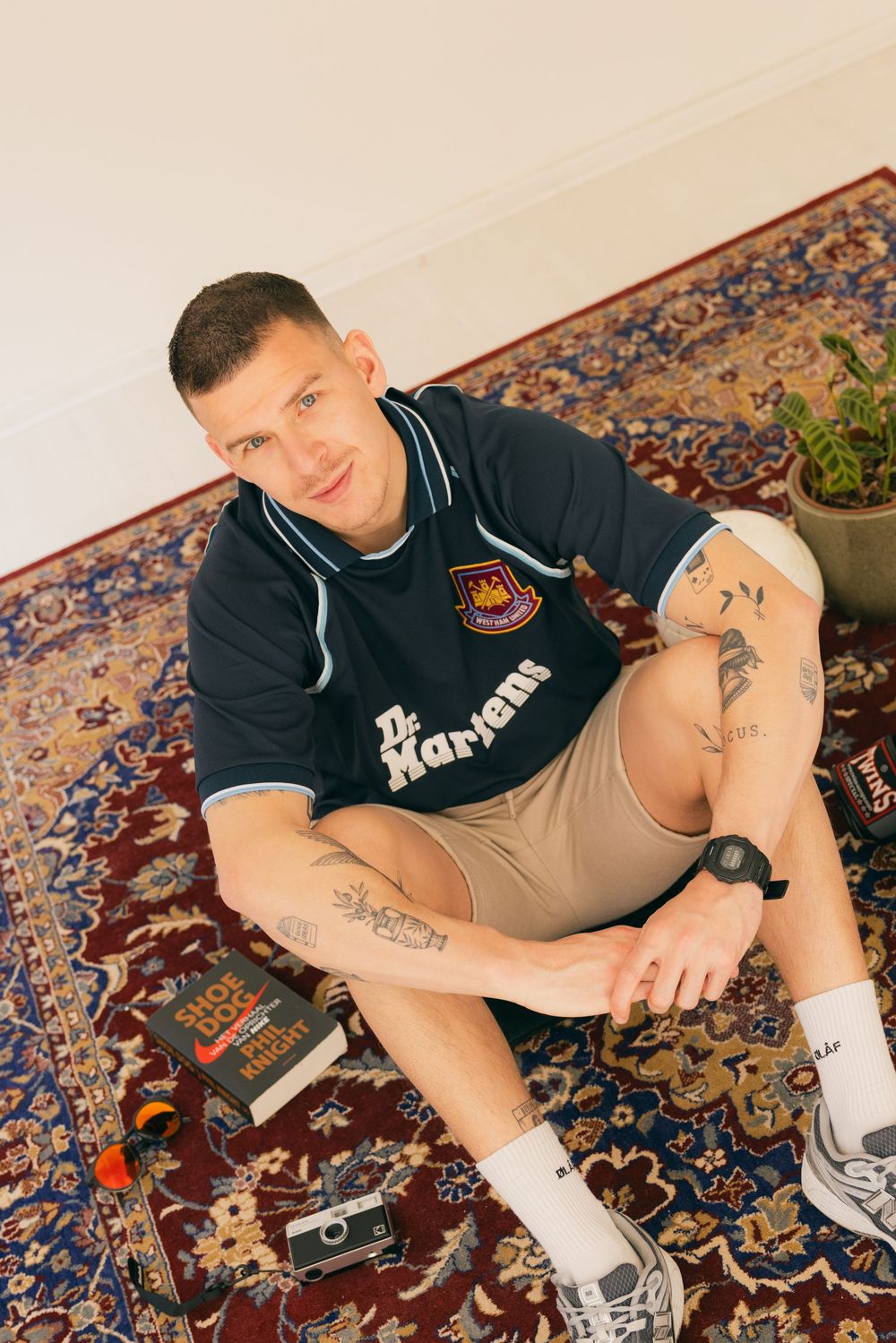 Young man with tattoos sitting on a patterned rug wearing a West Ham football shirt, khaki shorts, and white socks.