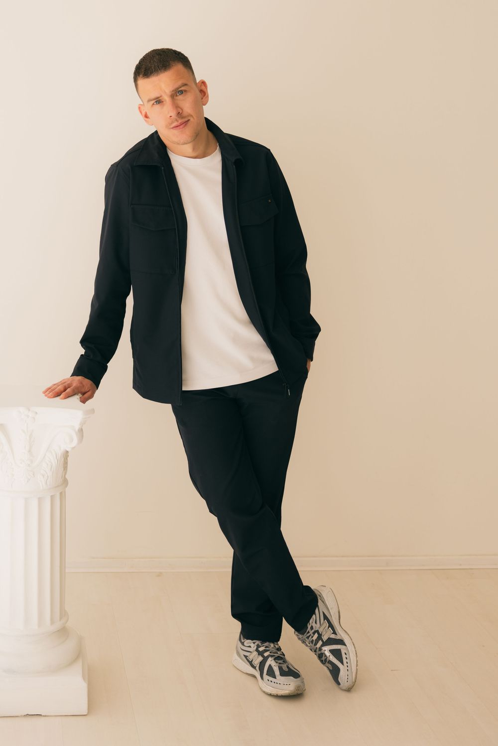 Man in black overshirt, trousers, white tee, and sneakers leaning on a white column pedestal.