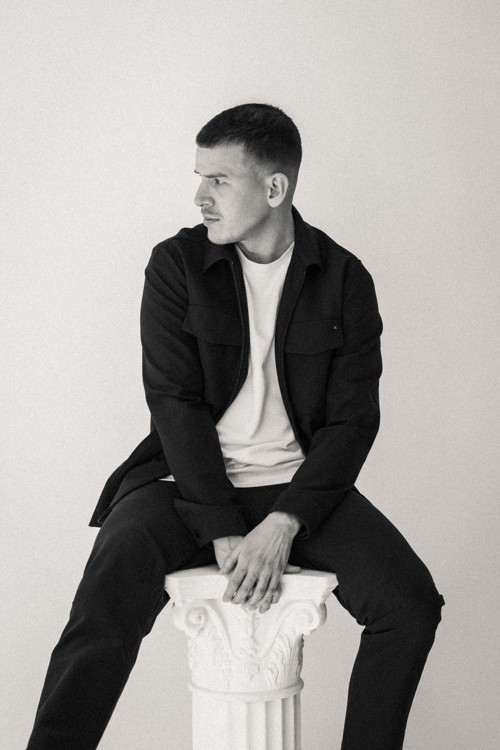 Black and white portrait of a young man in dark jacket sitting on a white classical column pedestal against light background.