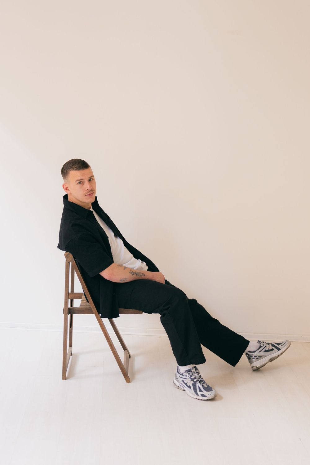Man in black suit and sneakers sitting relaxed on wooden chair against white background.