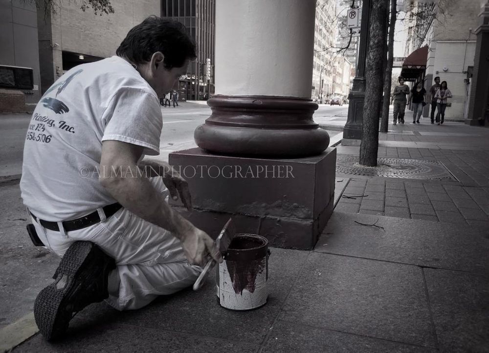 Someone kneeling to paint or maintain a concrete base on an urban sidewalk.