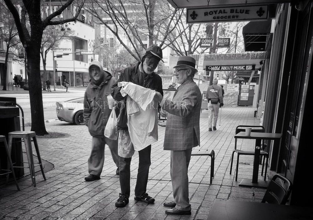 Three individuals stand on a city sidewalk reading a newspaper outside a storefront in a black and white photograph.
