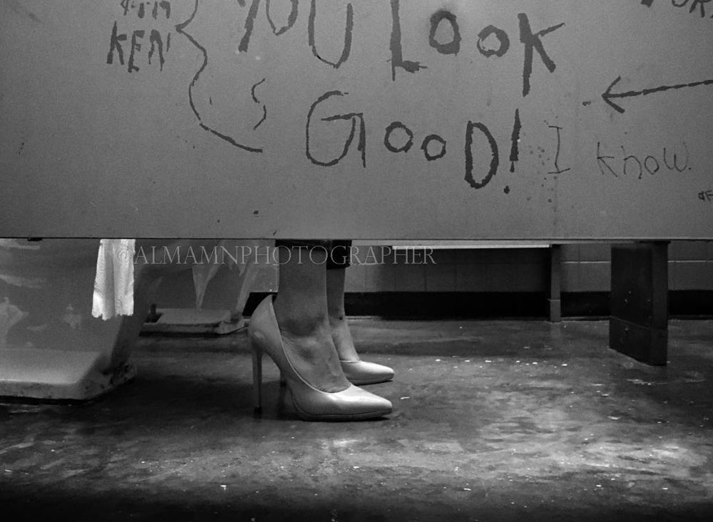 Black and white photo showing feet under a bathroom stall with graffiti visible above.