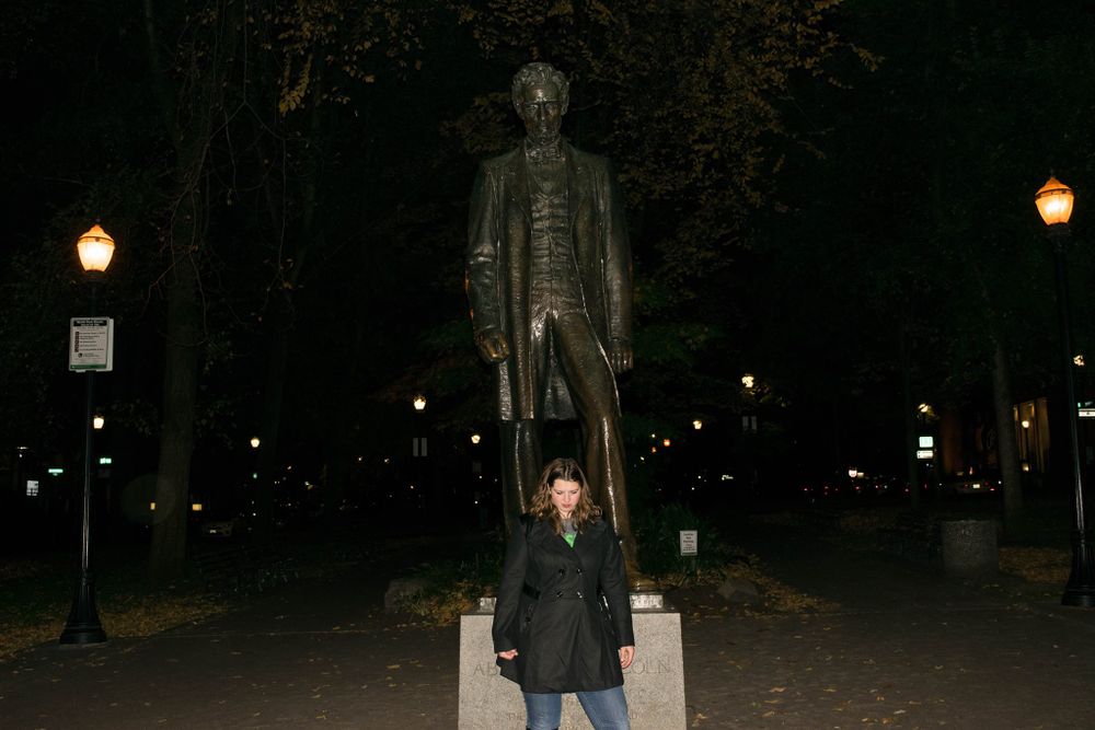 Tall bronze statue stands prominently in park at night with lamp posts glowing in background.