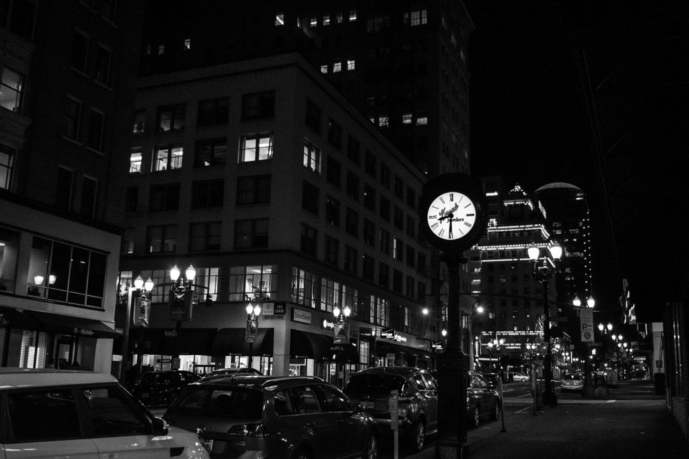 City street at night with illuminated clock and buildings in black and white.