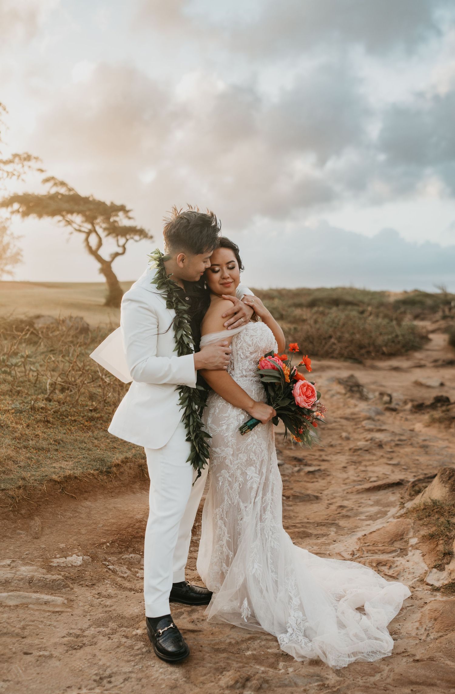 Sunset wedding portrait on rocky terrain with bride holding coral bouquet and groom wearing traditional lei.