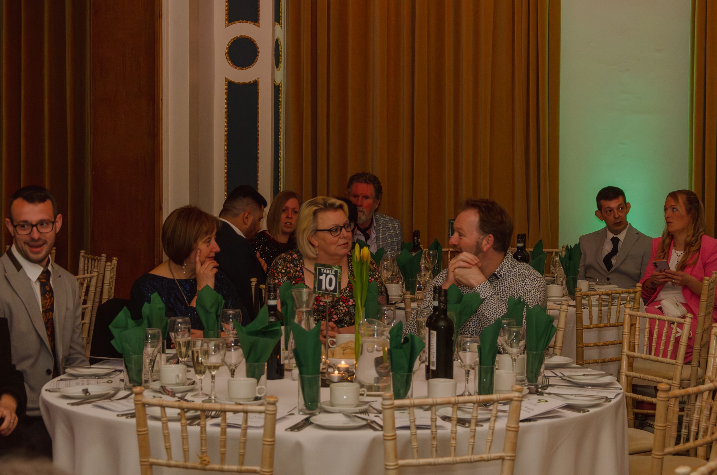 Guests seated at a formal dinner party with green lighting and wine bottles on the table.