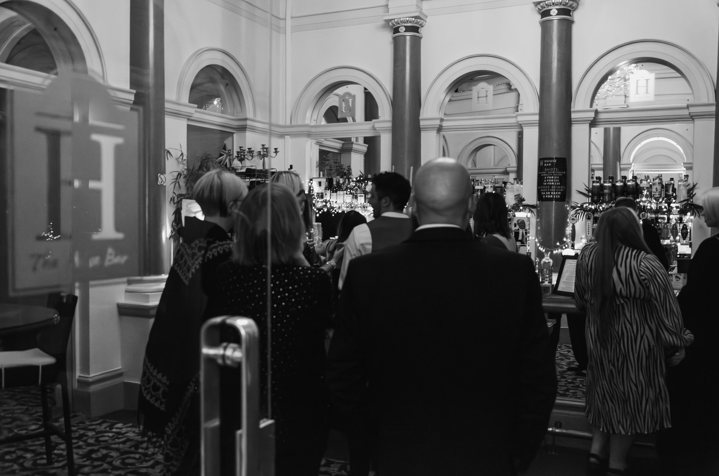 Black and white photo of guests mingling in an elegant venue with arched columns and architectural details.