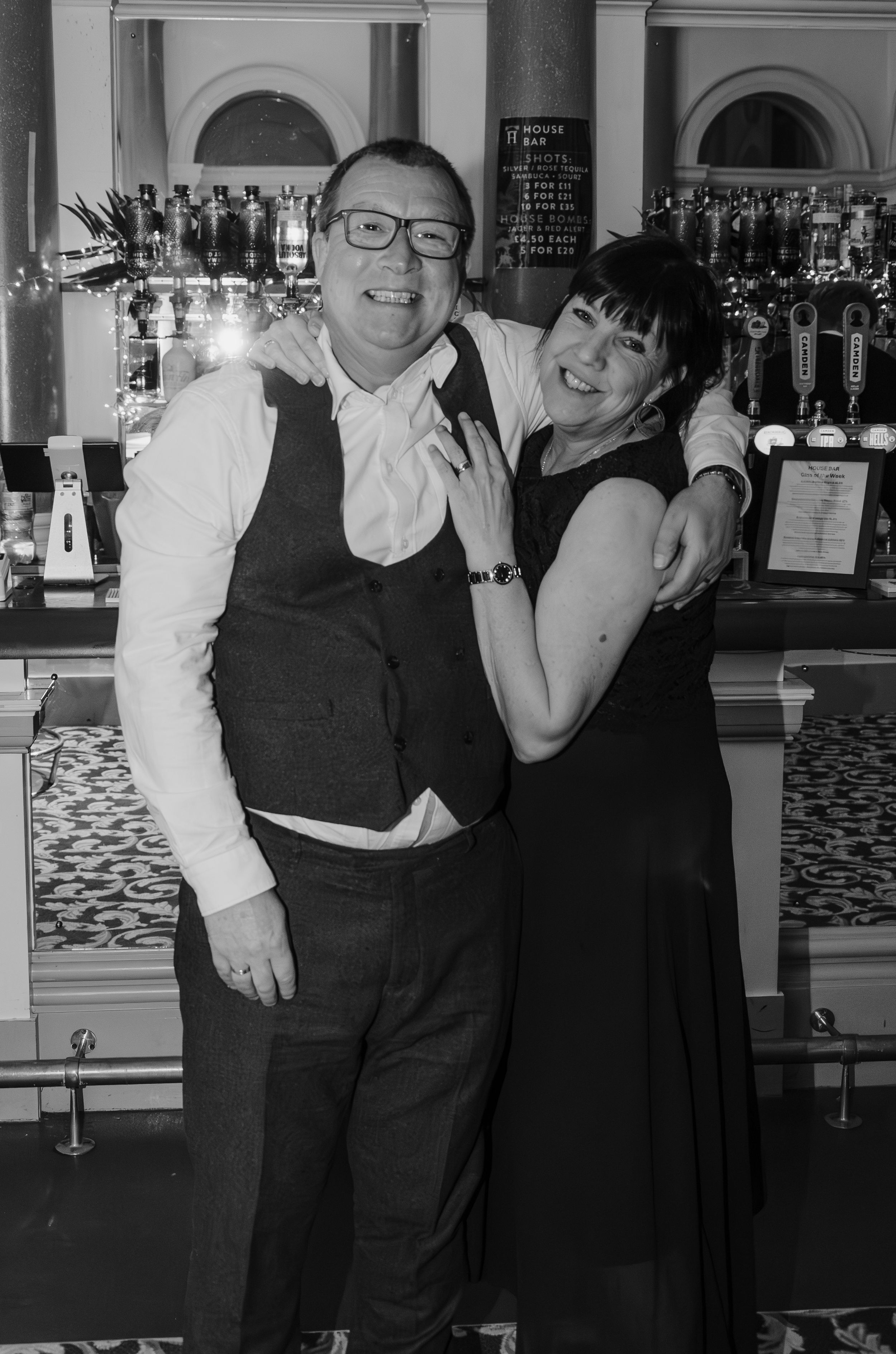 Two staff members pose together at an elegant bar with bottles displayed behind them.