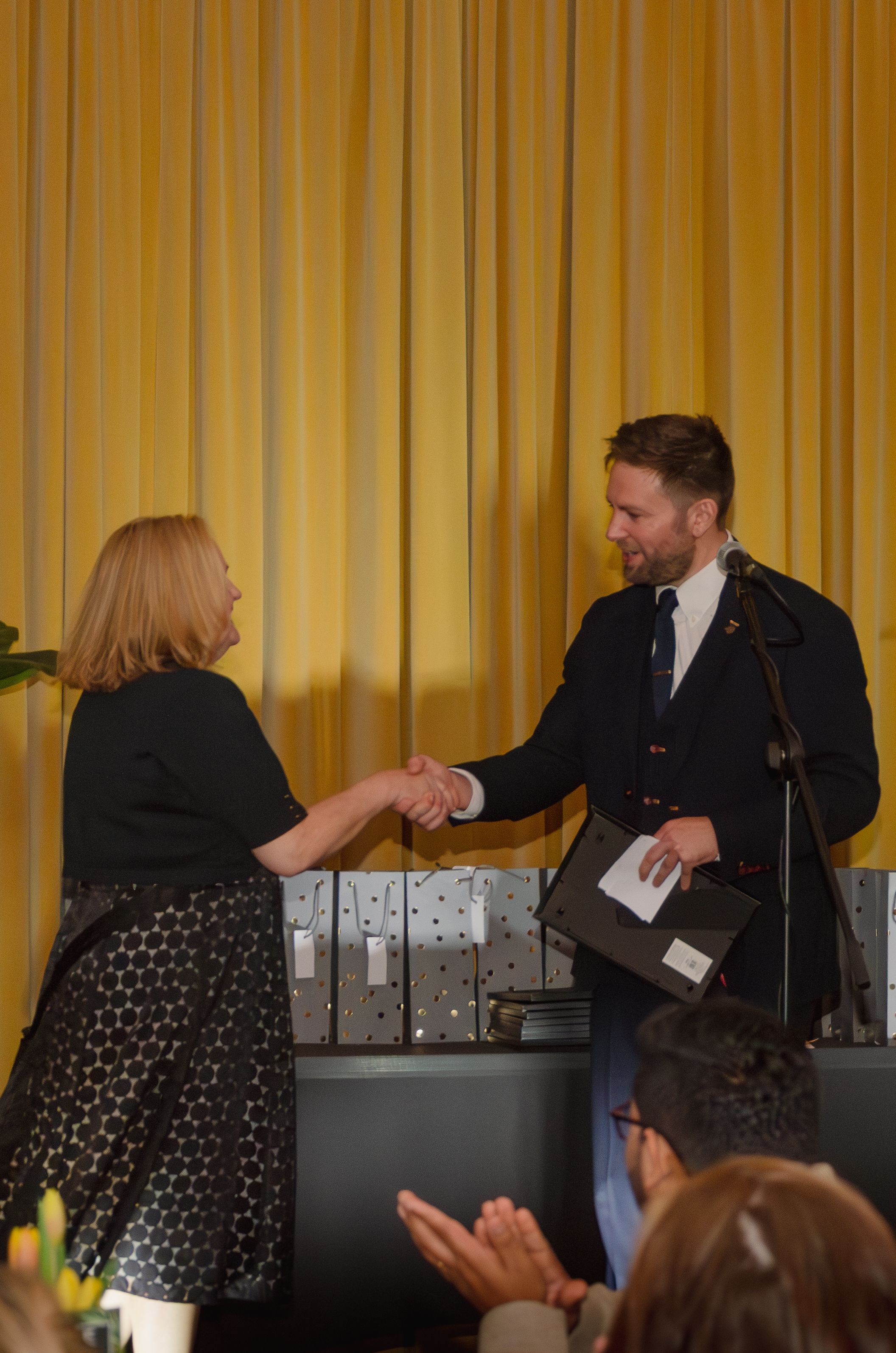 Two people shake hands on stage during an awards or recognition ceremony.