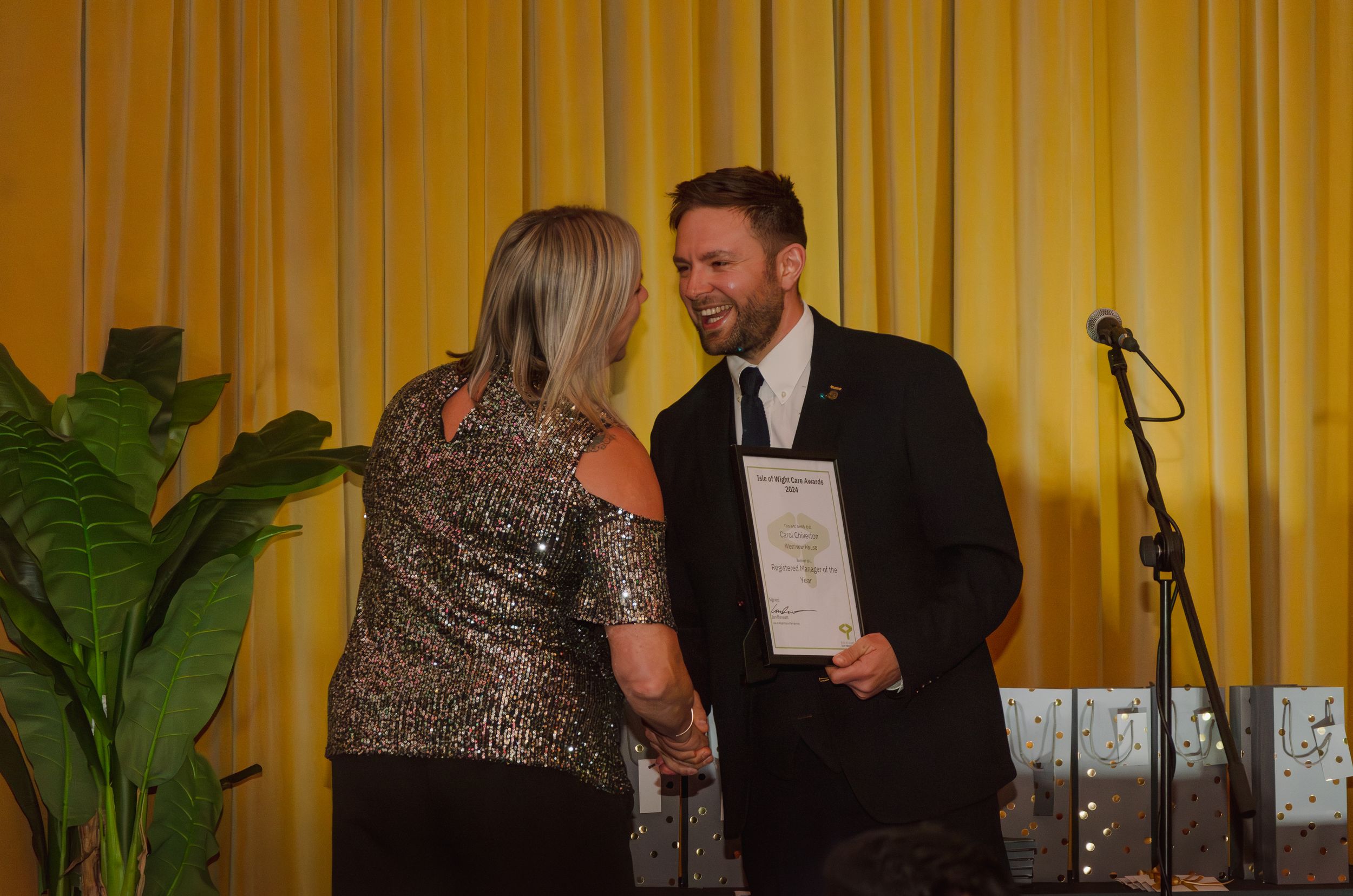 Two people exchange documents during an awards ceremony in front of a gold curtained backdrop.