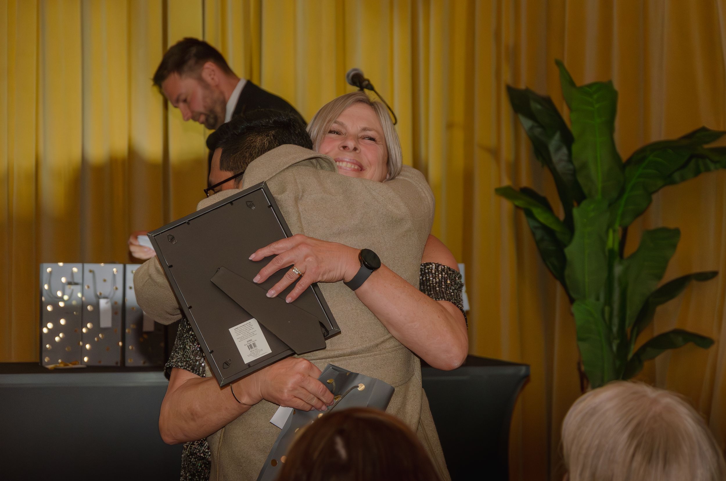 Two people sharing an emotional embrace during a celebratory event with yellow curtains in the background.