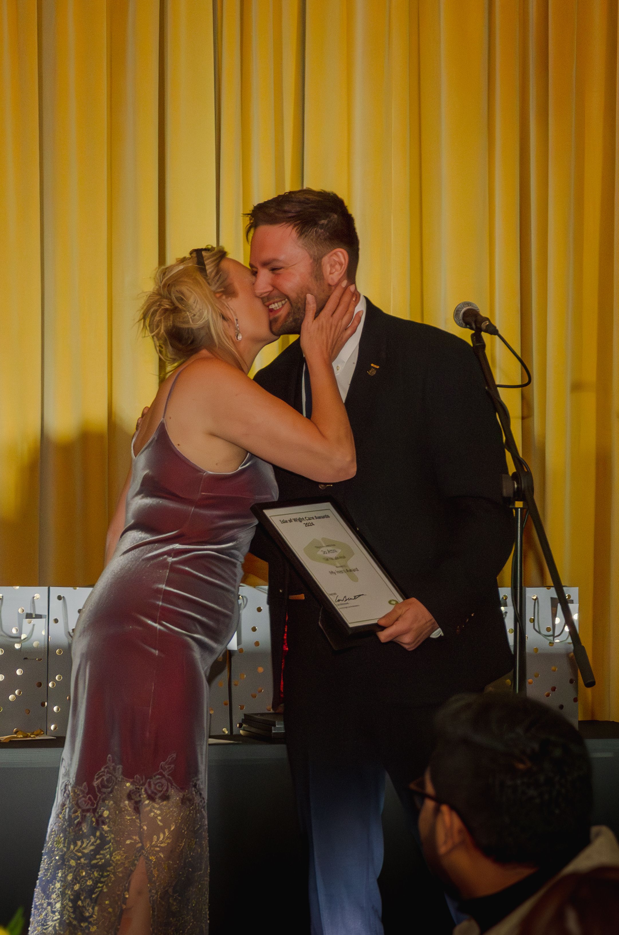 A romantic moment between a couple at a formal event with a microphone stand visible.