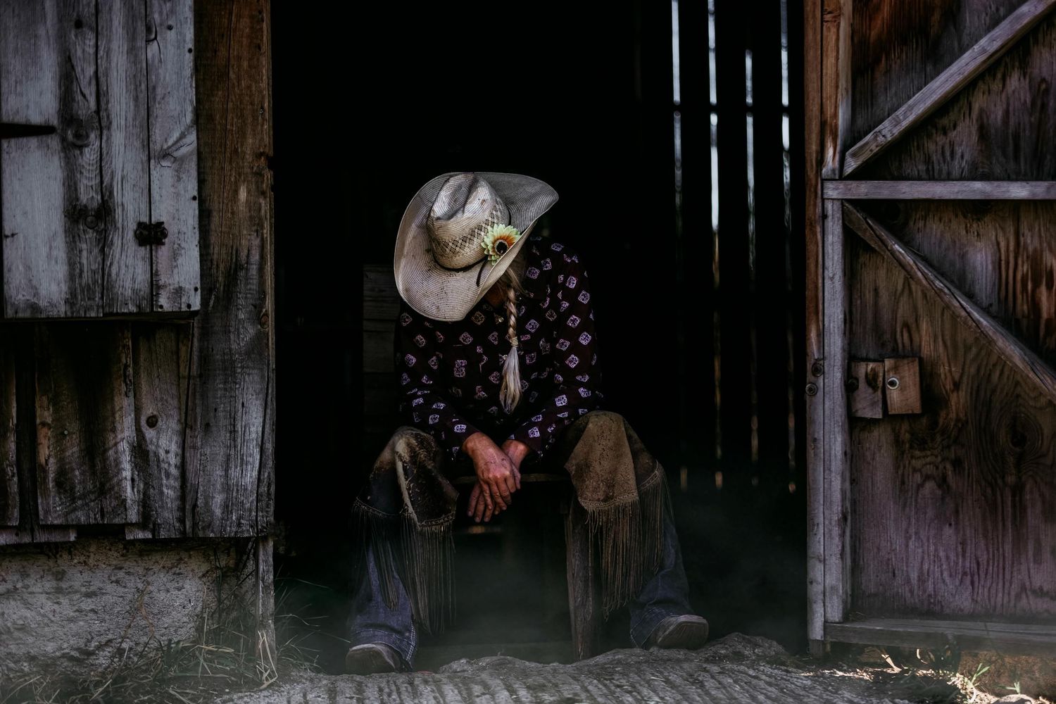 lady in cowboy hat sits contemplatively in darkened barn doorway.