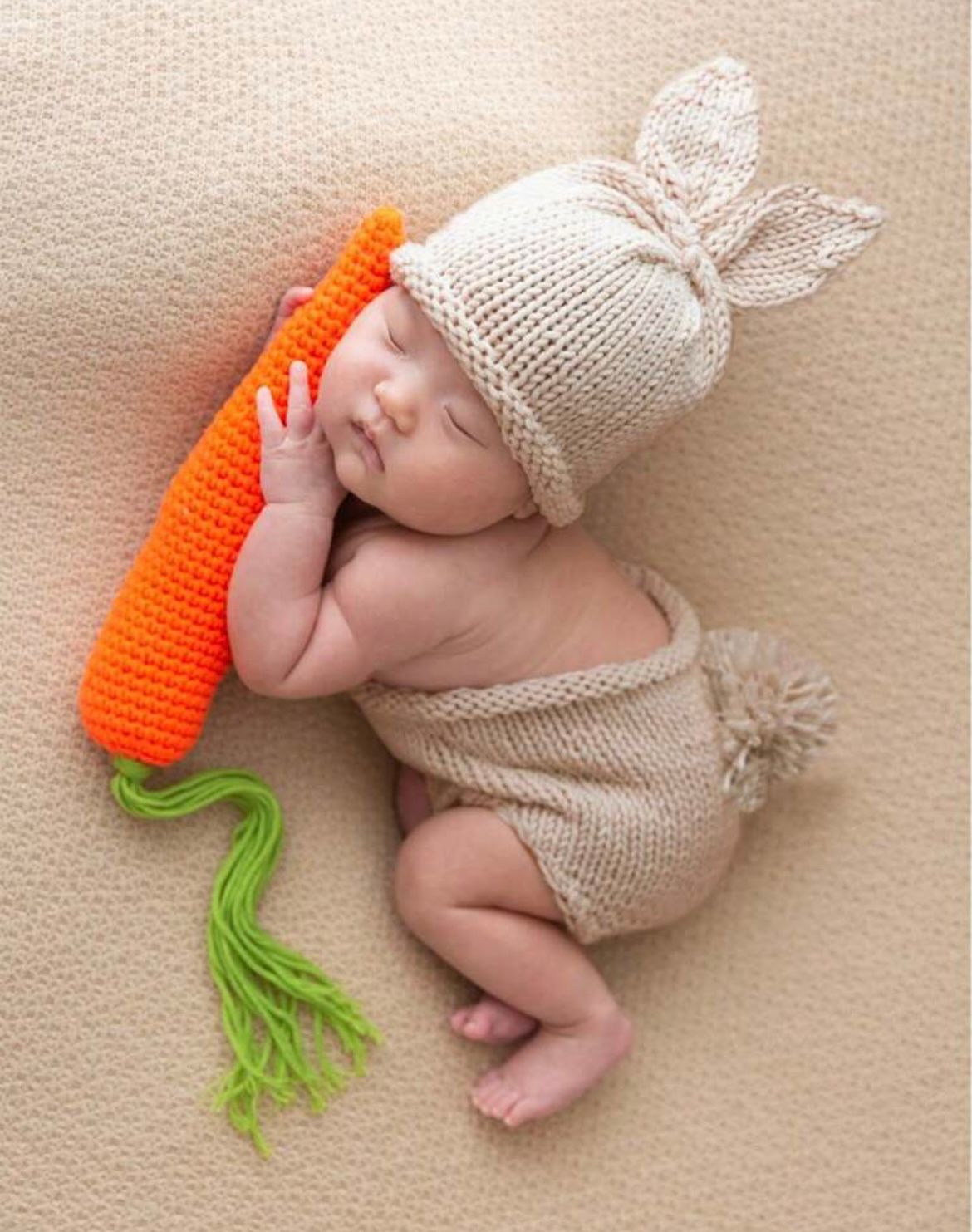 Sleeping newborn in white knit bunny outfit with orange carrot prop on textured background.
