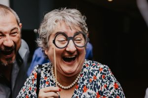 An elderly person in glasses and a floral print top laughs joyfully with genuine emotion.