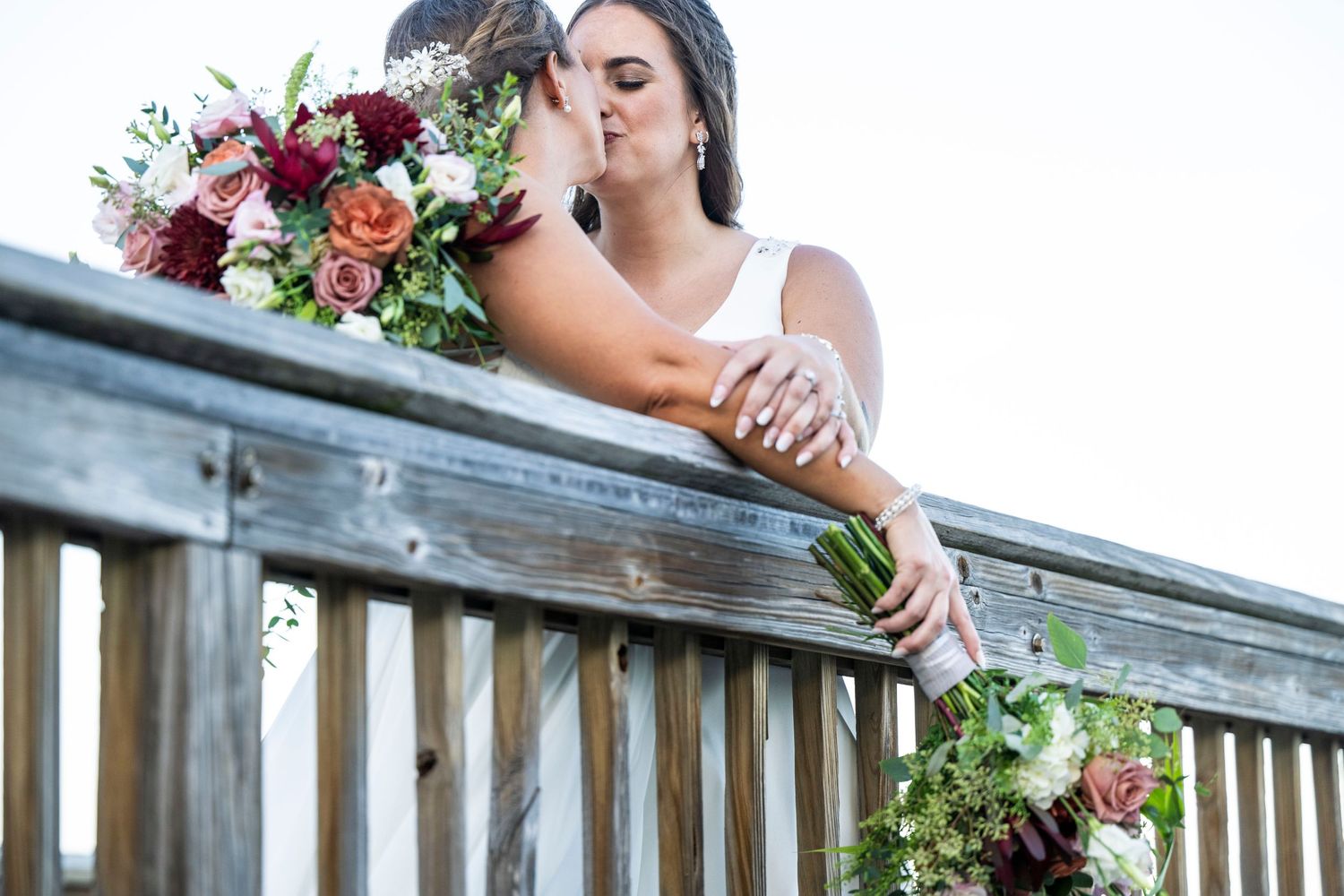 Bride holding vibrant red and white bouquet while leaning on wooden deck railing.